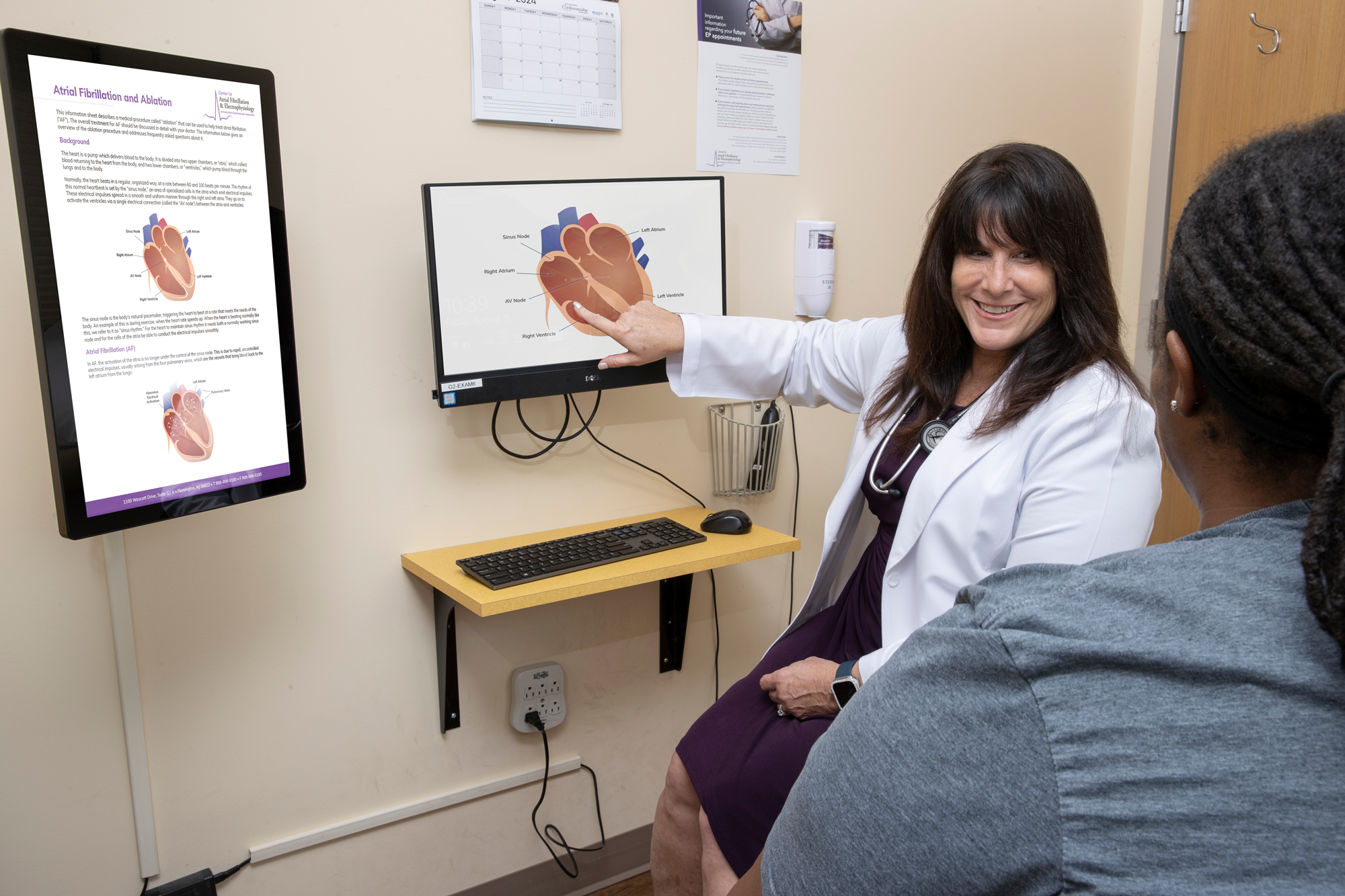 A brown-haired doctor points at a diagram of a heart displayed on a computer monitor with a smile on her face. A patient is watching her intently.